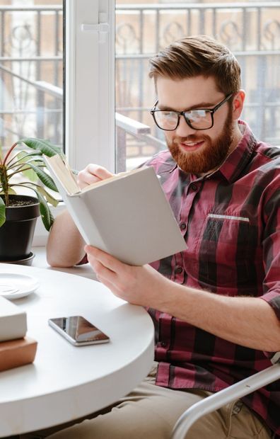Man with glasses and beard reads a book at a table near a window.