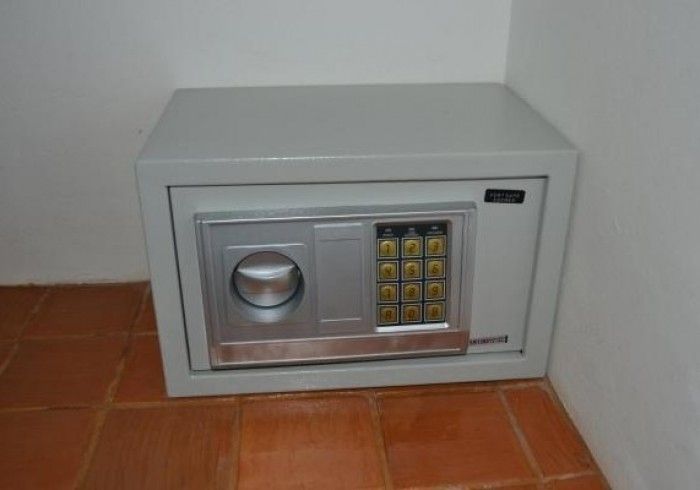 Cofre cinza com teclado numérico e mostrador, instalado em um canto sobre um piso de azulejos.