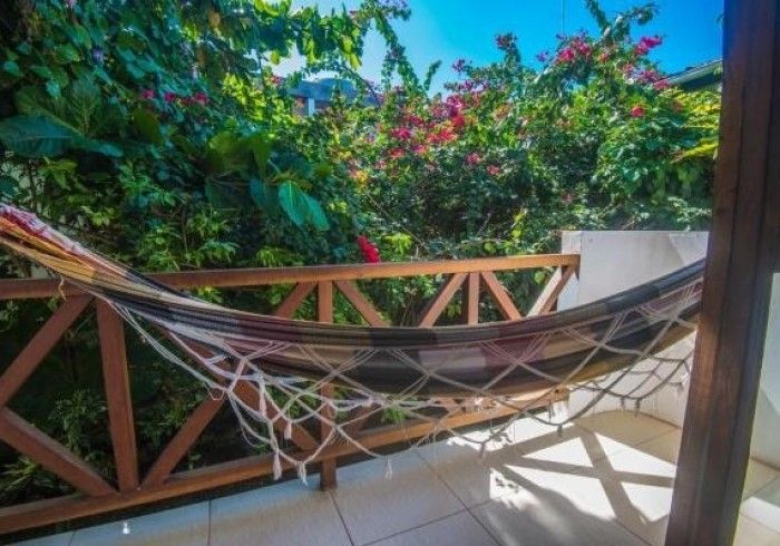 Rede numa varanda com vista para plantas floridas exuberantes em tons de verde e rosa, sob um céu azul.