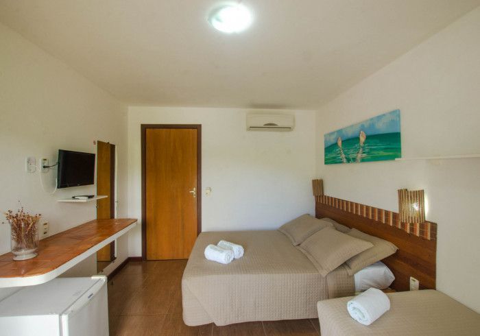 Quarto com cama, TV pequena, escrivaninha, porta de madeira e um quadro com uma paisagem de praia.