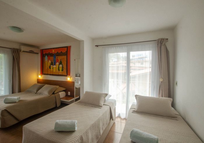 Quarto com três camas: duas de solteiro e uma de casal. Roupa de cama bege, obras de arte e janela com cortinas.