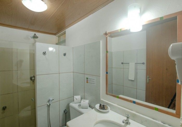 Banheiro com paredes revestidas de azulejos brancos, chuveiro, vaso sanitário, espelho e pia. Teto e porta de madeira marrom.