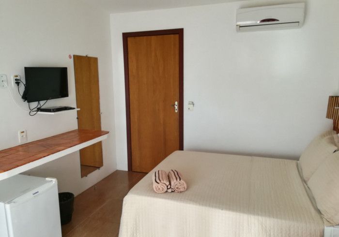 Quarto de hotel pequeno com cama, TV, escrivaninha, frigobar e porta; paredes brancas, porta e escrivaninha de madeira.