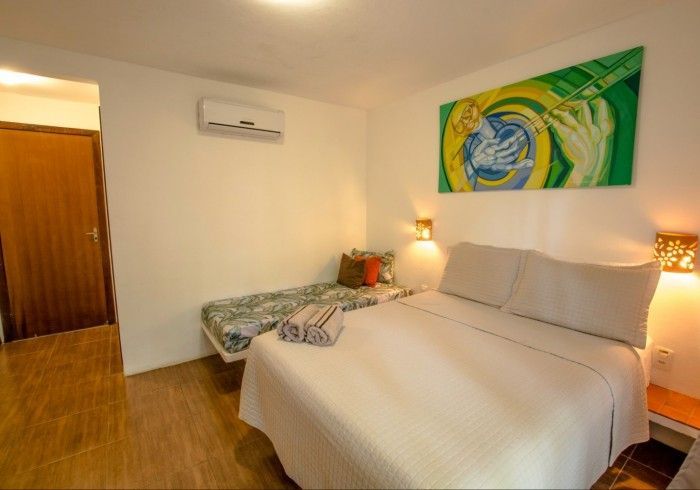 Quarto com cama de casal, cama de solteiro, obras de arte, ar condicionado, piso de madeira e porta.