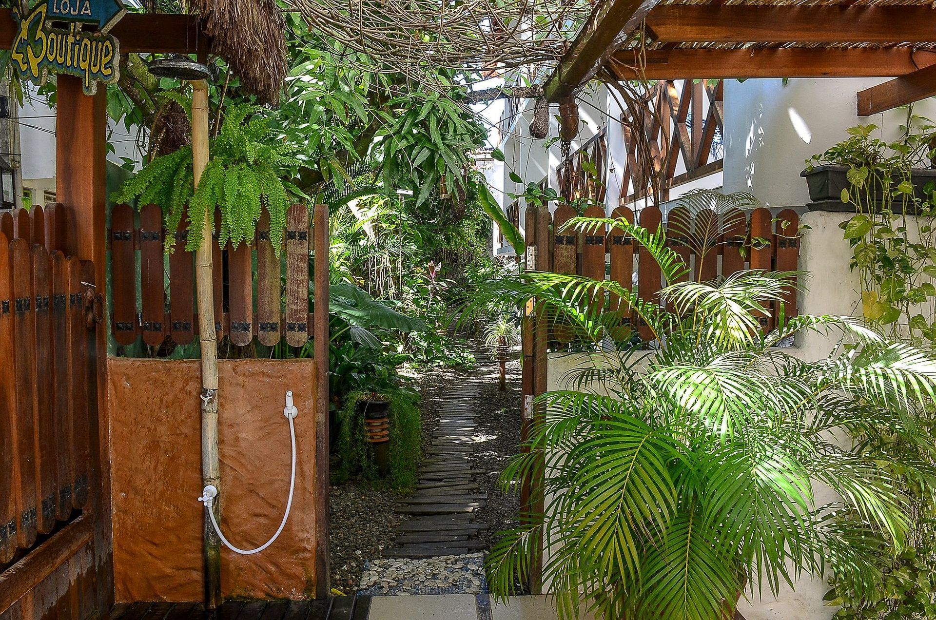 Passarela externa exuberante e sombreada com portão de madeira, caminho de pedra e vegetação abundante.