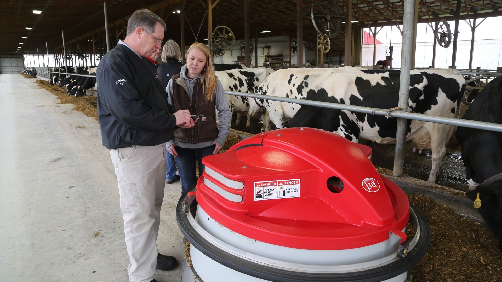 Two people standing beside a red robotic feed pusher in a dairy barn.