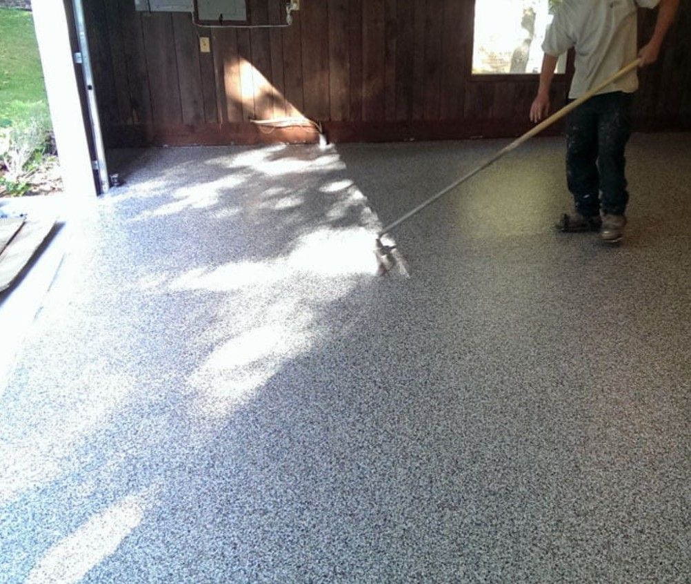 A person uses a long-handled tool to apply a speckled, gray epoxy finish to a garage floor.
