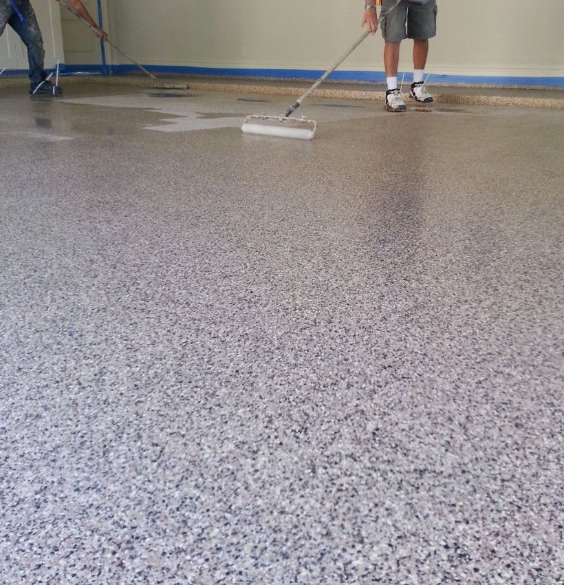 Two people use rollers to apply a speckled, glossy coating to a garage floor.