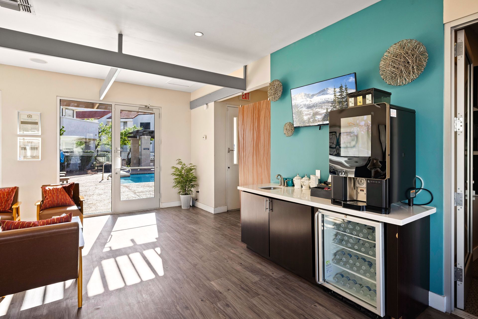 Lounge area with coffee machine, mini-fridge, teal wall, and view of a pool through glass doors. Experience modern Tempe, Arizona apartments at Talavera Apartments today.