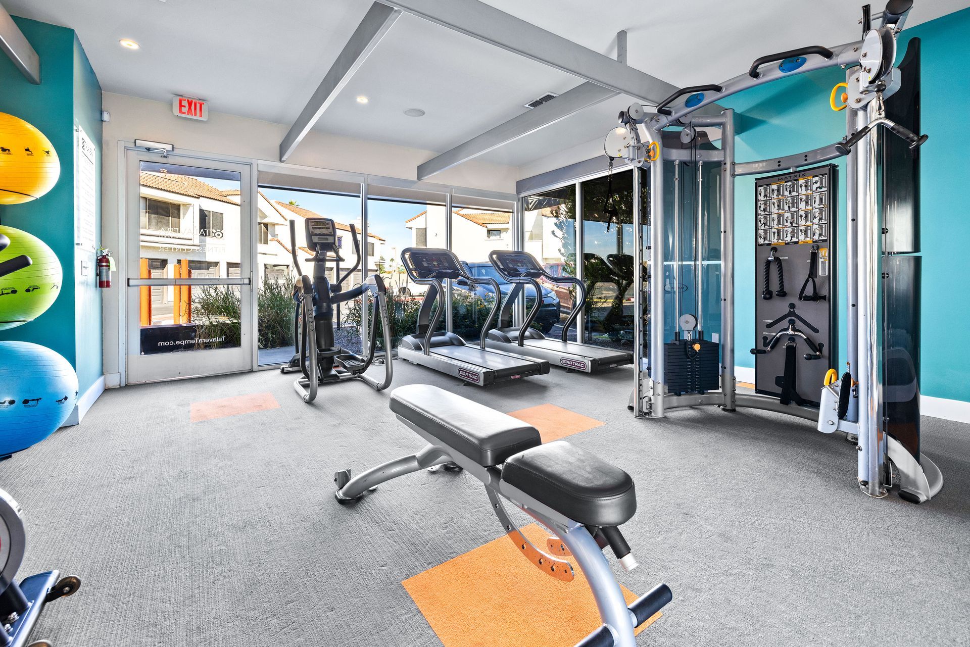 Gym with weight bench, treadmills, elliptical, and cable machine; teal and orange accents. Talavera Apartments highlights pet-friendly apartments in Tempe AZ—see the details.