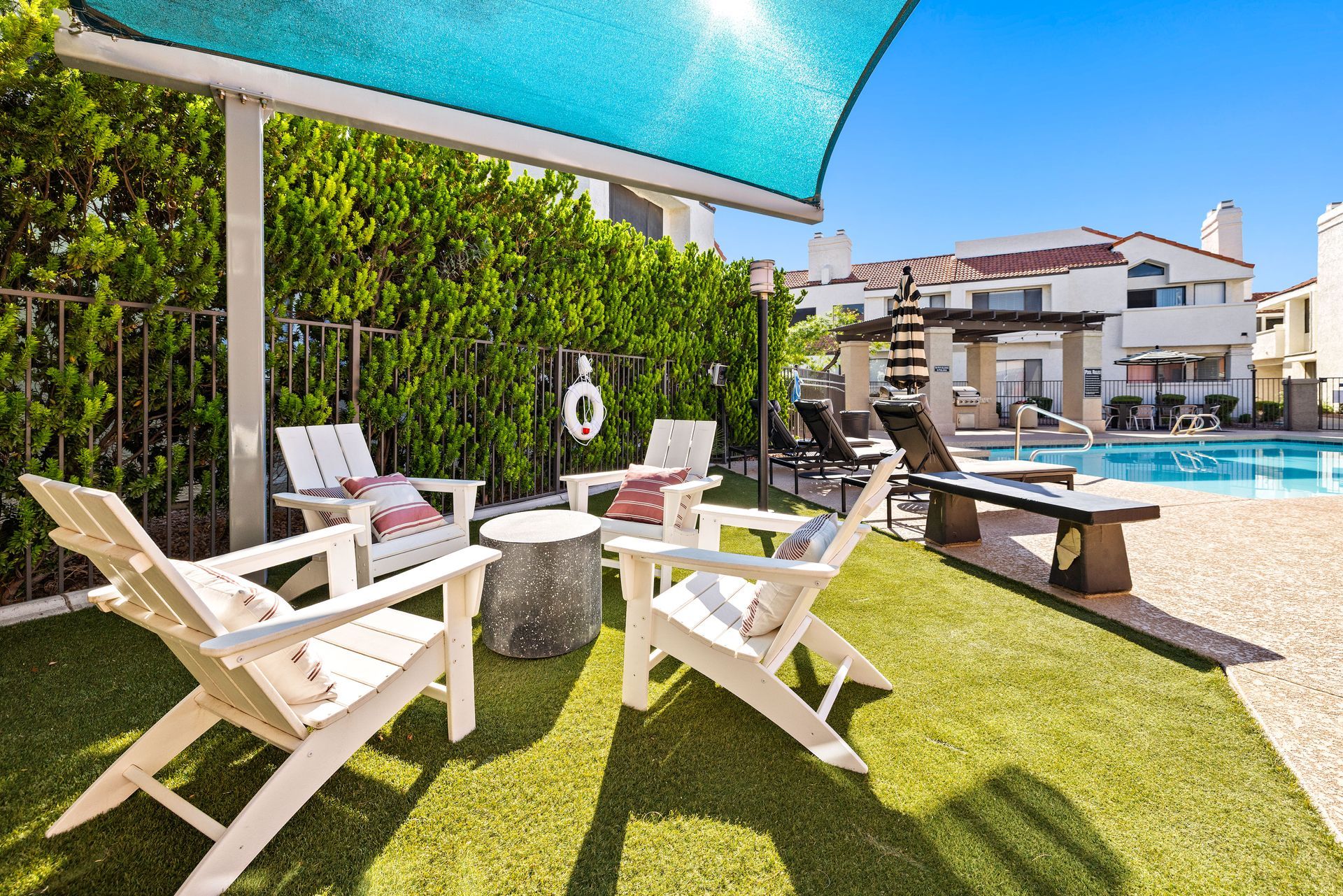 Outdoor seating area by a pool with white chairs, shade cover, and landscaping.