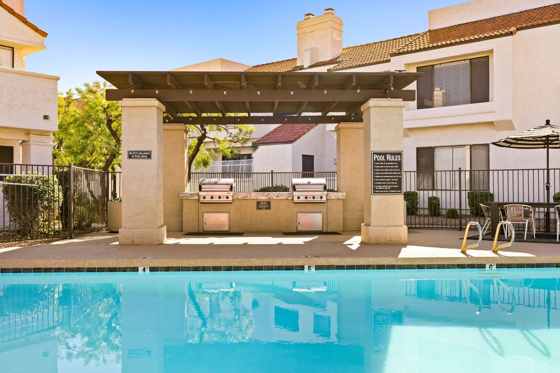 Poolside grilling area with two grills under a pergola, adjacent to a pool and apartment buildings. Talavera Apartments features well-designed Tempe AZ rentals—check them out.