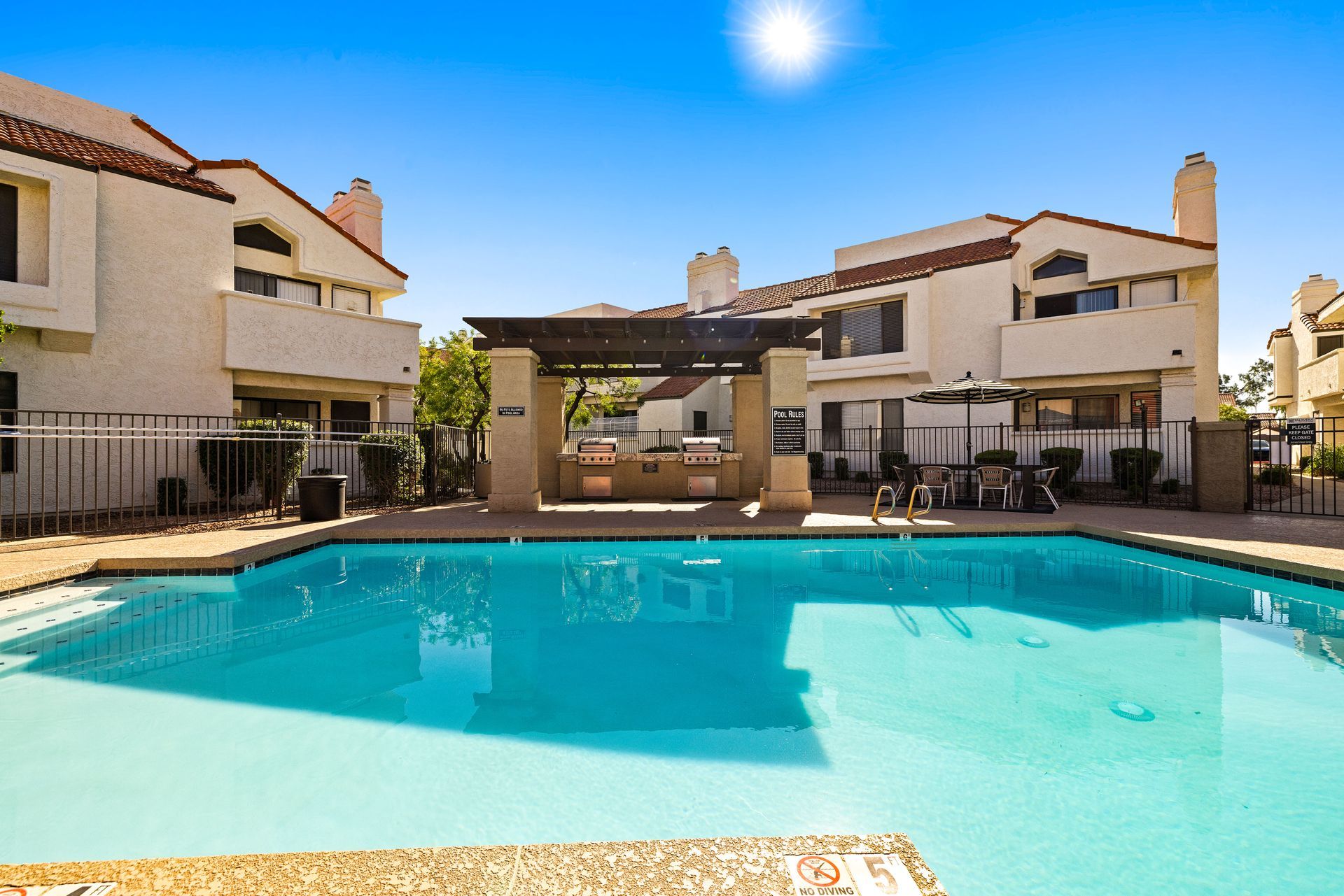 Pool in front of beige multi-story building, blue sky.  Outdoor grill and tables visible. Tour contemporary 2 bedroom apartments in Tempe AZ at Talavera Apartments.