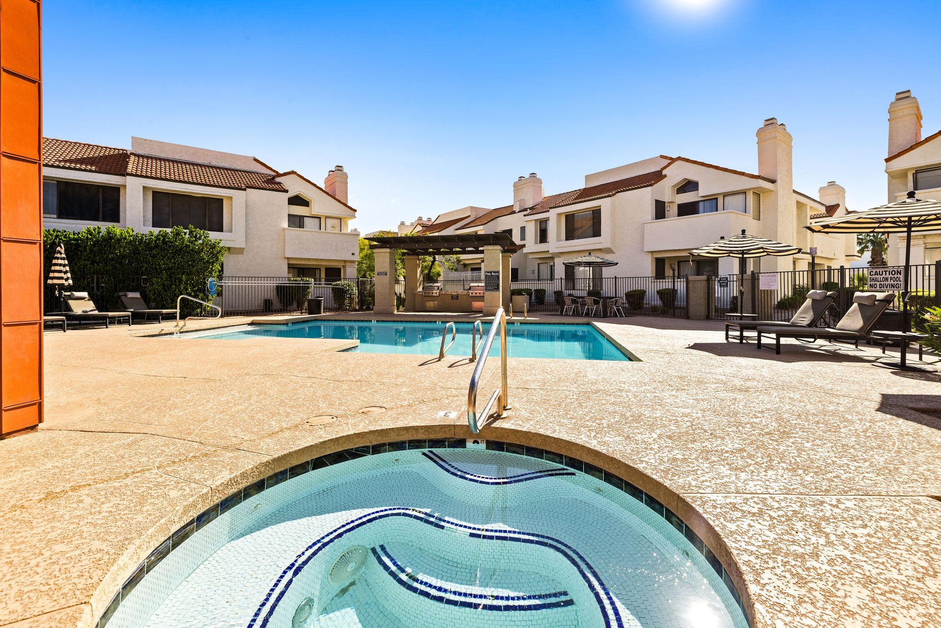 Pool area with jacuzzi in foreground, buildings in background under a blue sky. Talavera Apartments brings you closer with downtown Tempe apartments—learn more.