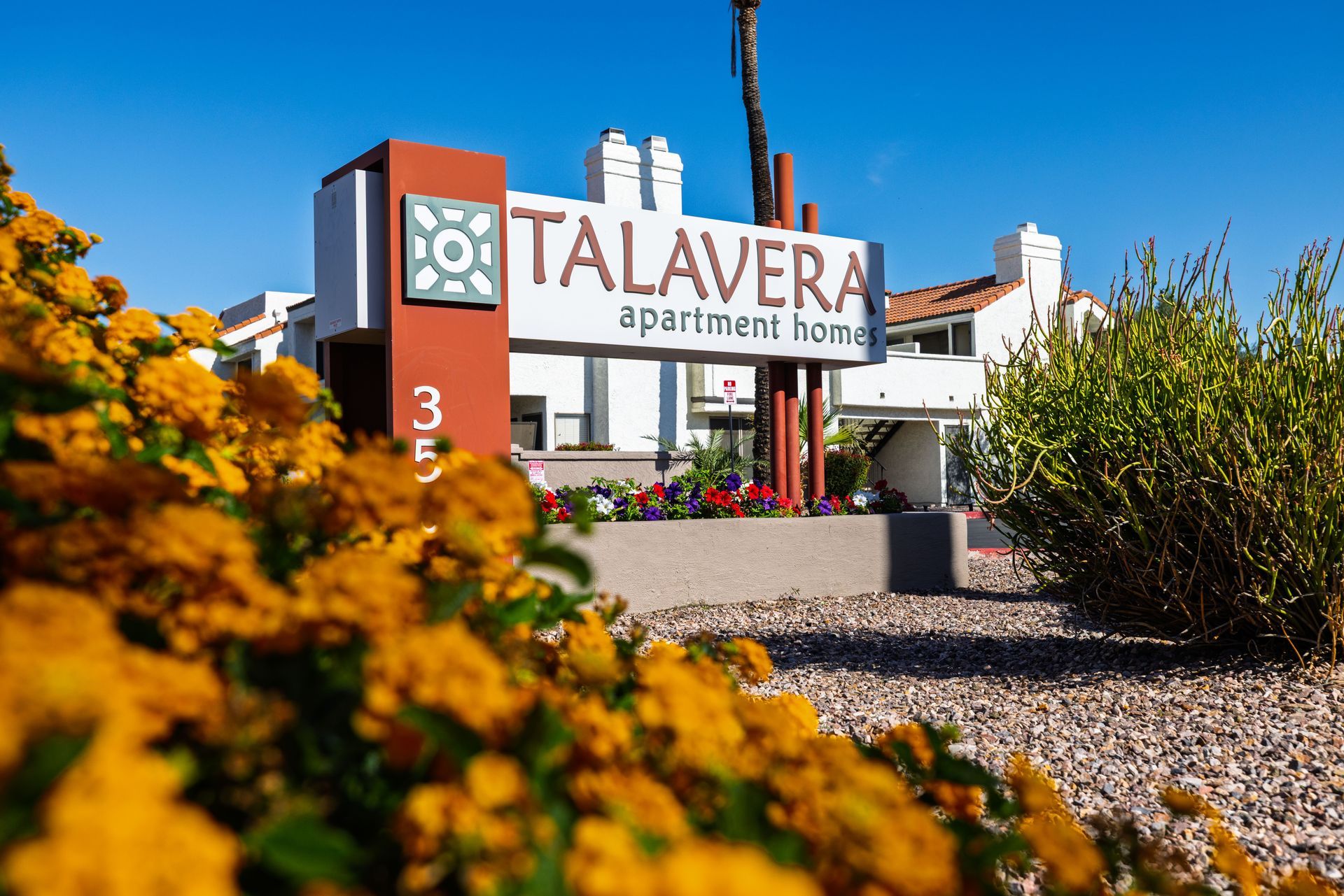 Talavera apartment complex sign with white text, surrounded by orange flowers and green plants under a blue sky.