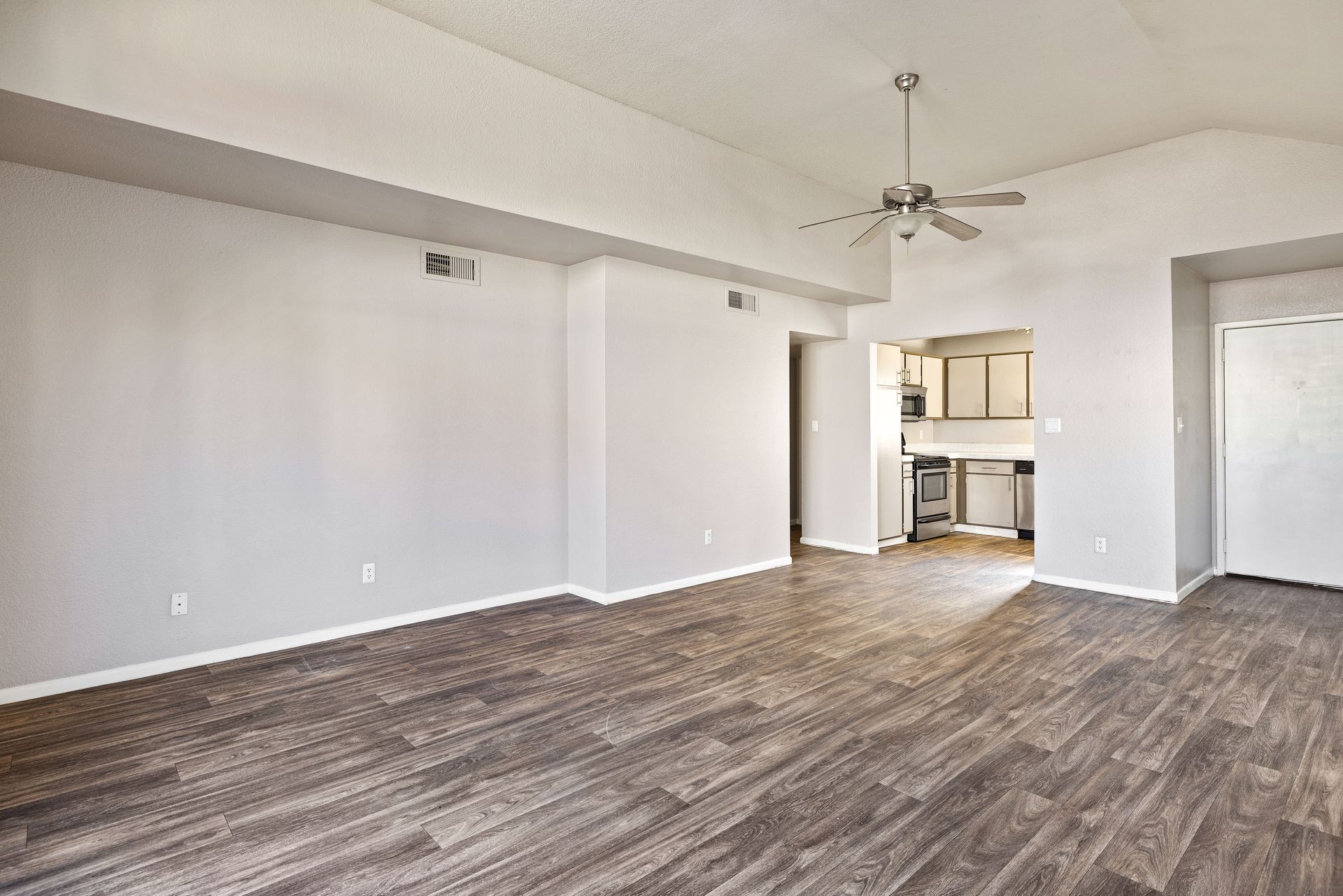 Empty living room with wood-look flooring, pale gray walls, ceiling fan, and glimpse of kitchen. Enjoy life in a pet-friendly community at Talavera Apartments—learn more today.