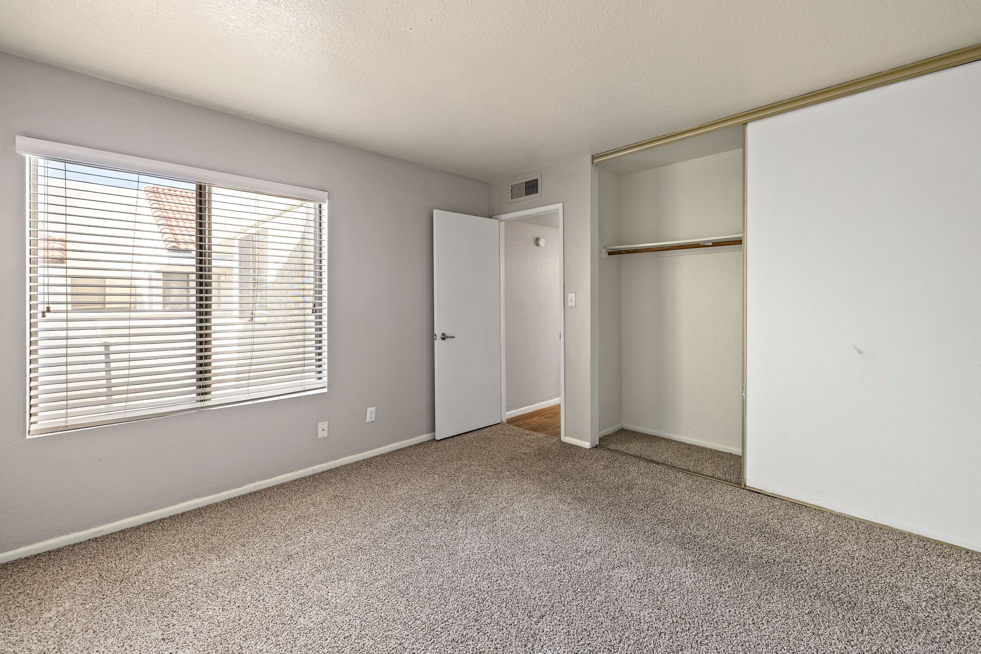 Empty bedroom with gray walls, carpet, window, open closet, and a closed door. Explore contemporary Tempe, Arizona apartments at Talavera Apartments today.