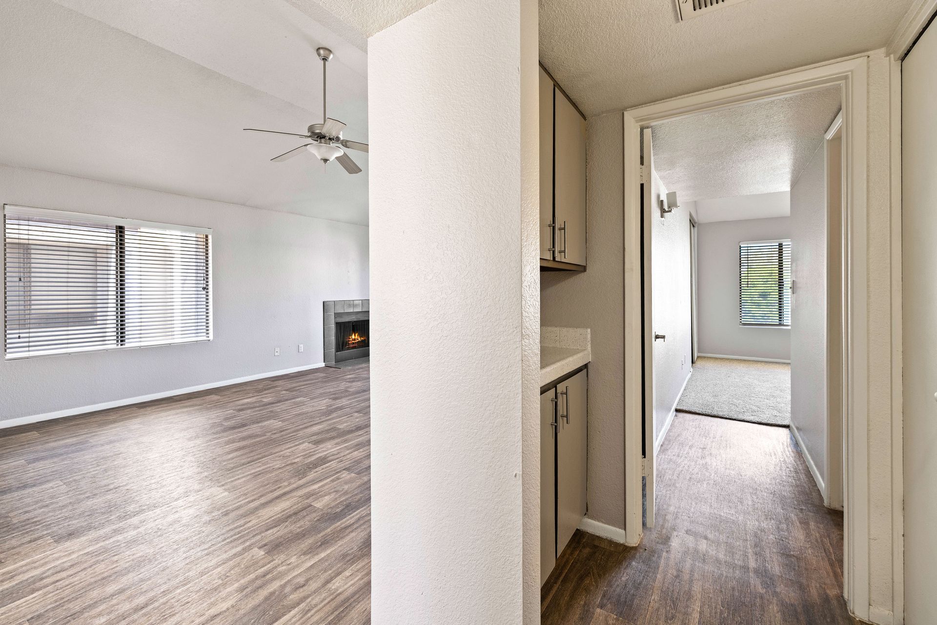 Interior view of a living space and hallway with wood-look flooring, fireplace, and window with blinds. Talavera Apartments offers comfortable Tempe AZ rentals—find your next home.