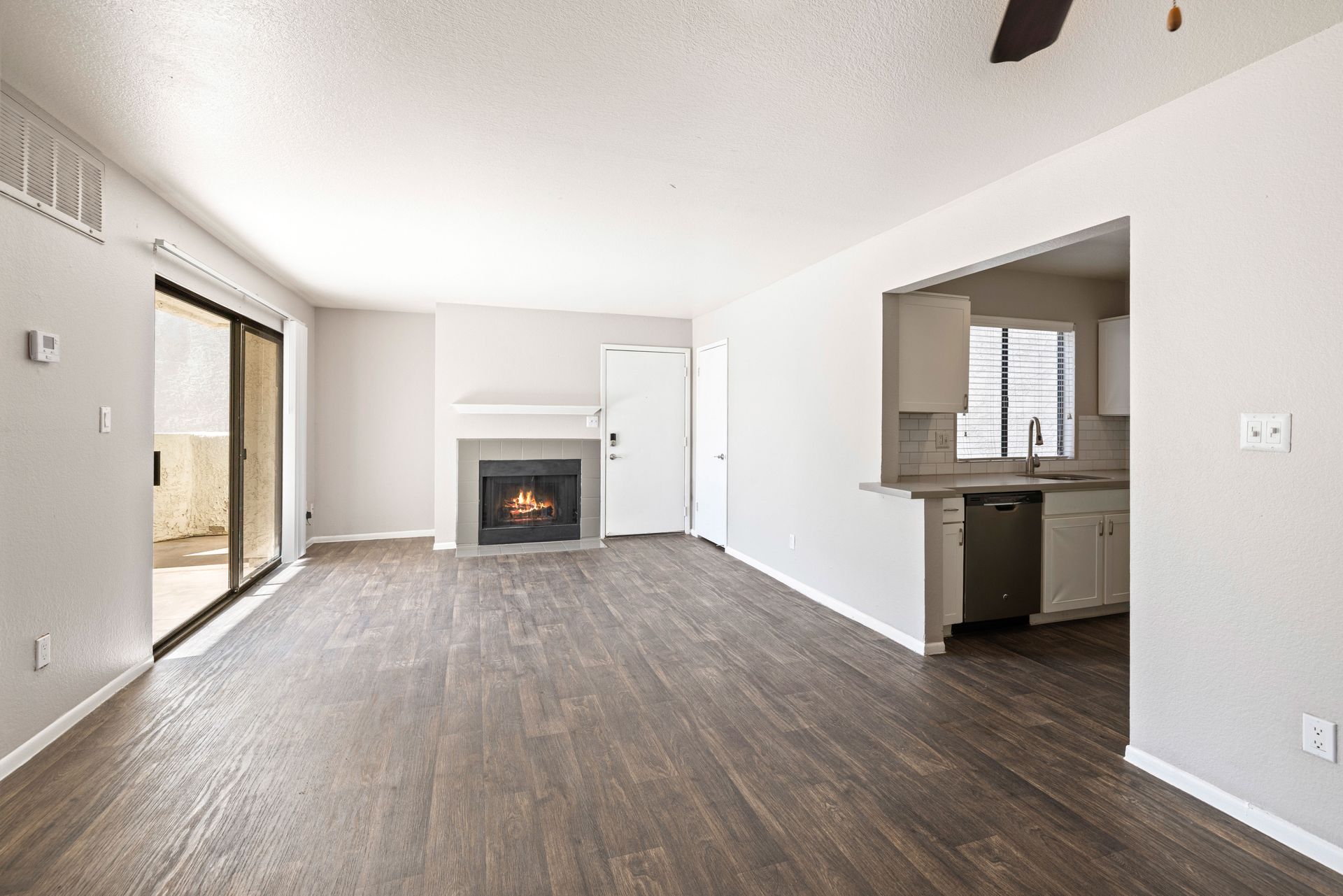Empty living room with fireplace, sliding door to patio, and view of kitchen. Gray walls and dark wood floors. Tour apartment homes in Tempe AZ at Talavera Apartments and picture your lifestyle.