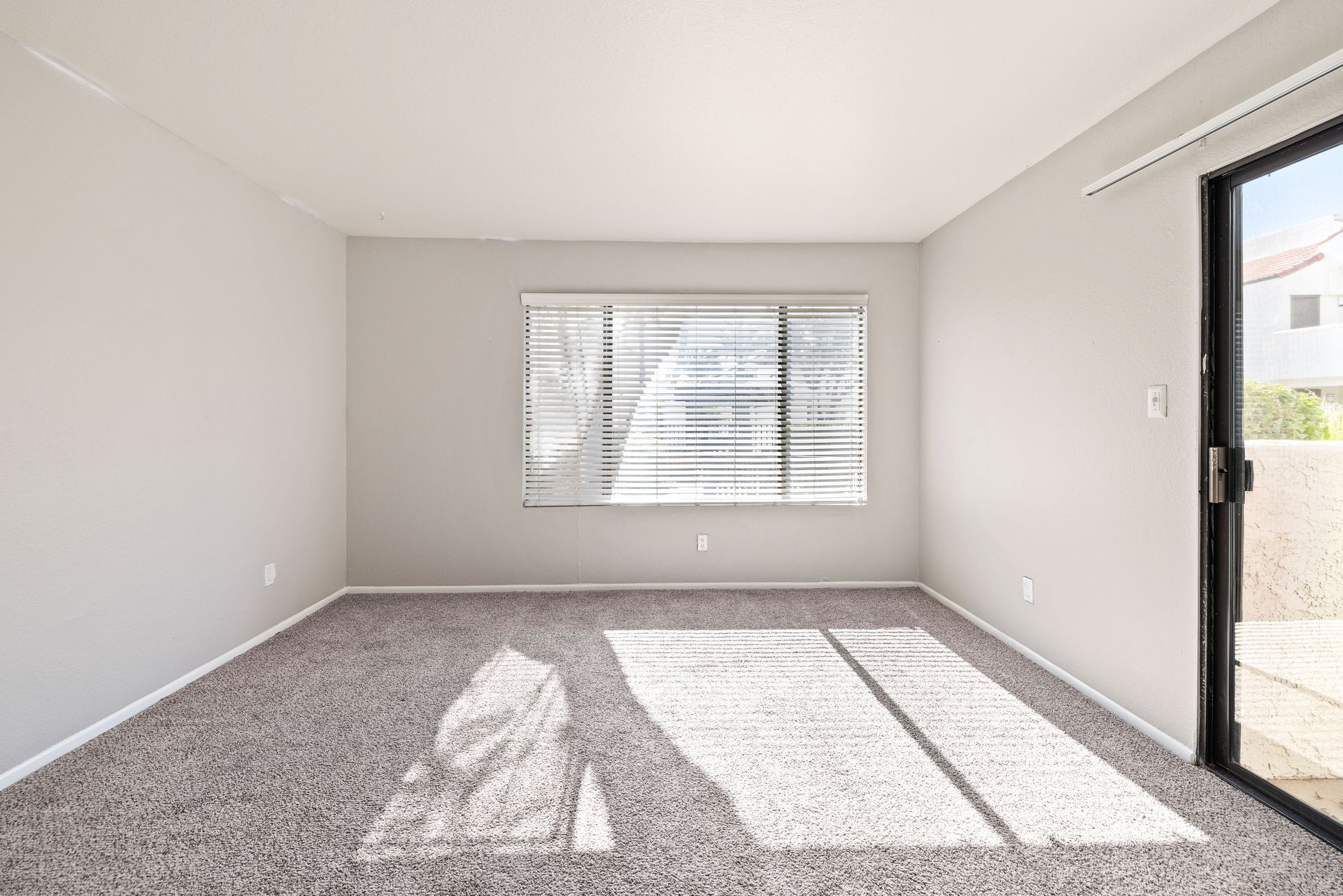 Empty room with light gray walls, carpet, window with blinds, and sliding glass door. Tour beautifully designed 2 bedroom apartments in Tempe AZ at Talavera Apartments.