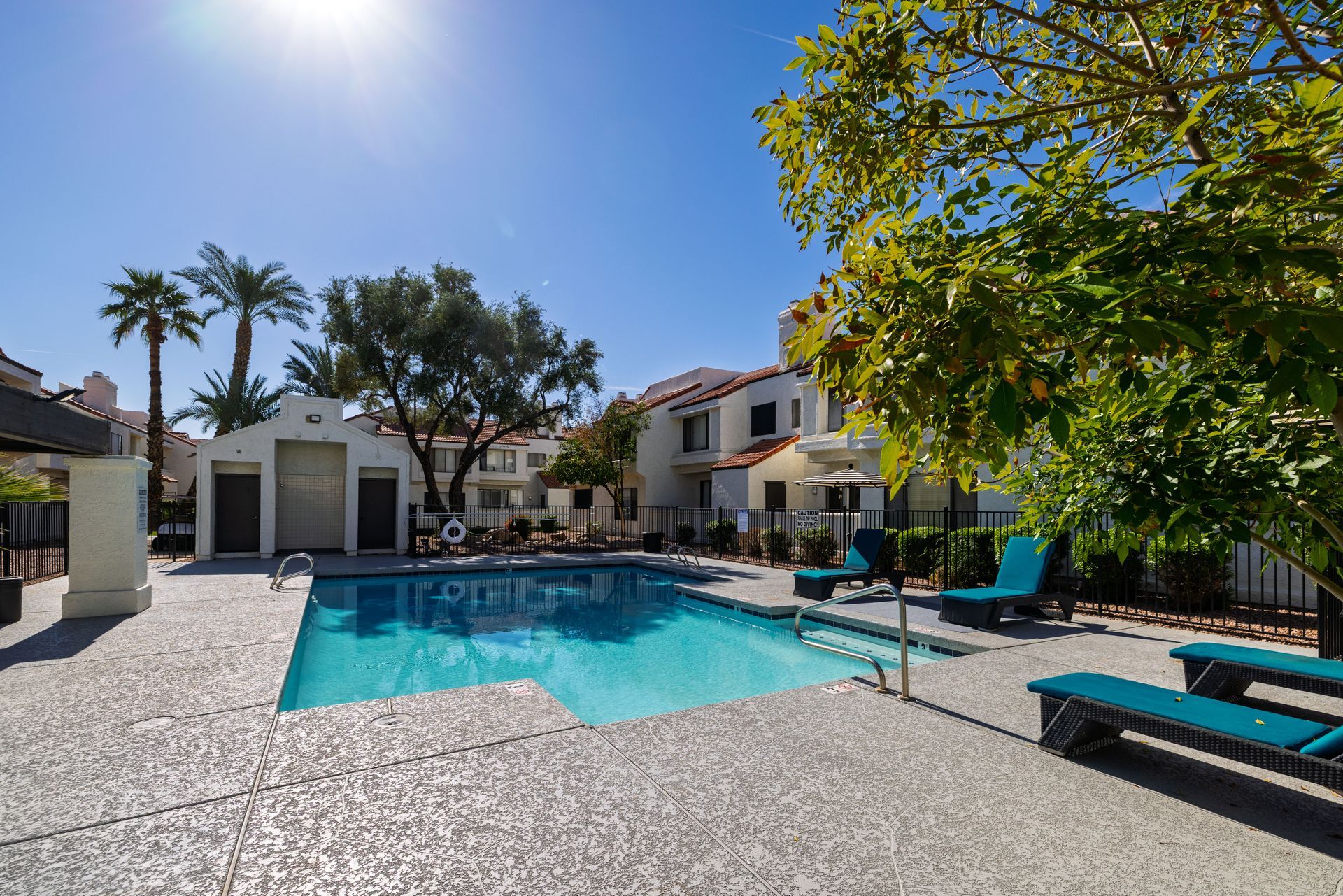 Pool area with turquoise water, palm trees, and two-story buildings under a bright blue sky.