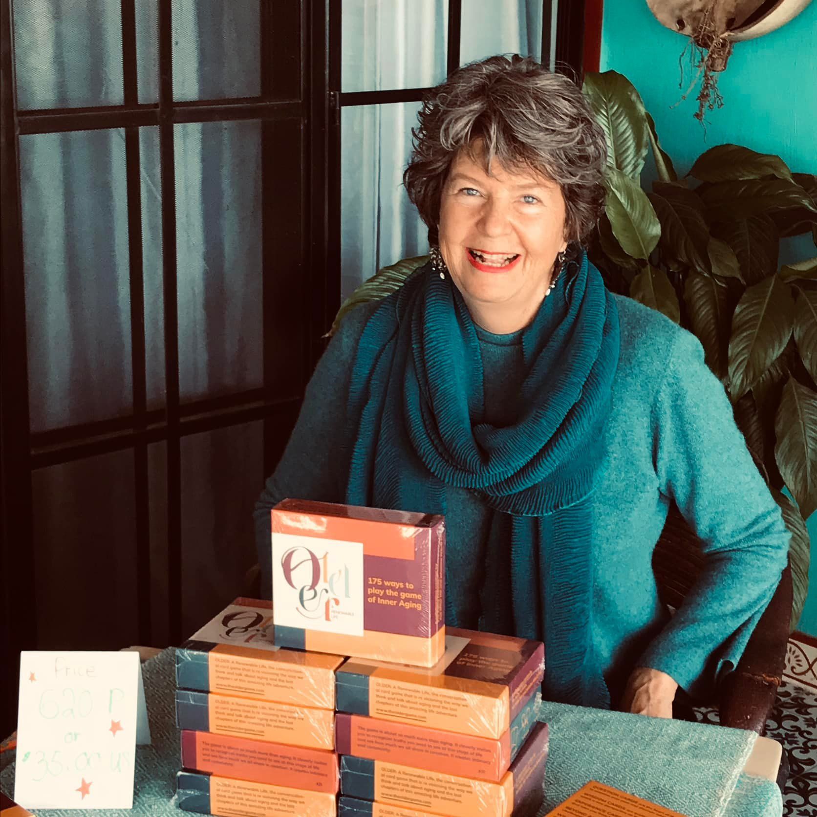 Woman with gray hair smiles, surrounded by books. She wears a teal sweater. Sunlight streams through the window.