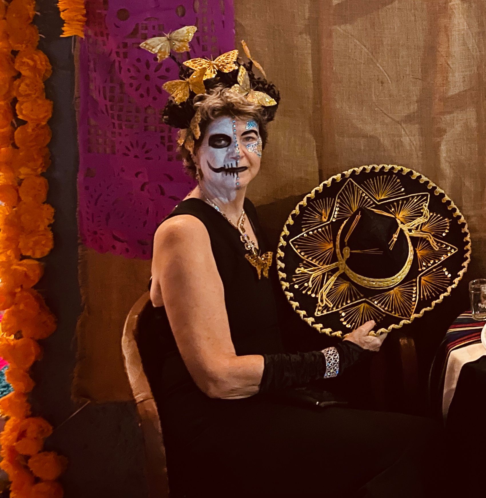 Woman in Day of the Dead makeup holds a sombrero, sits in front of a colorful backdrop, and has butterfly hair decor.