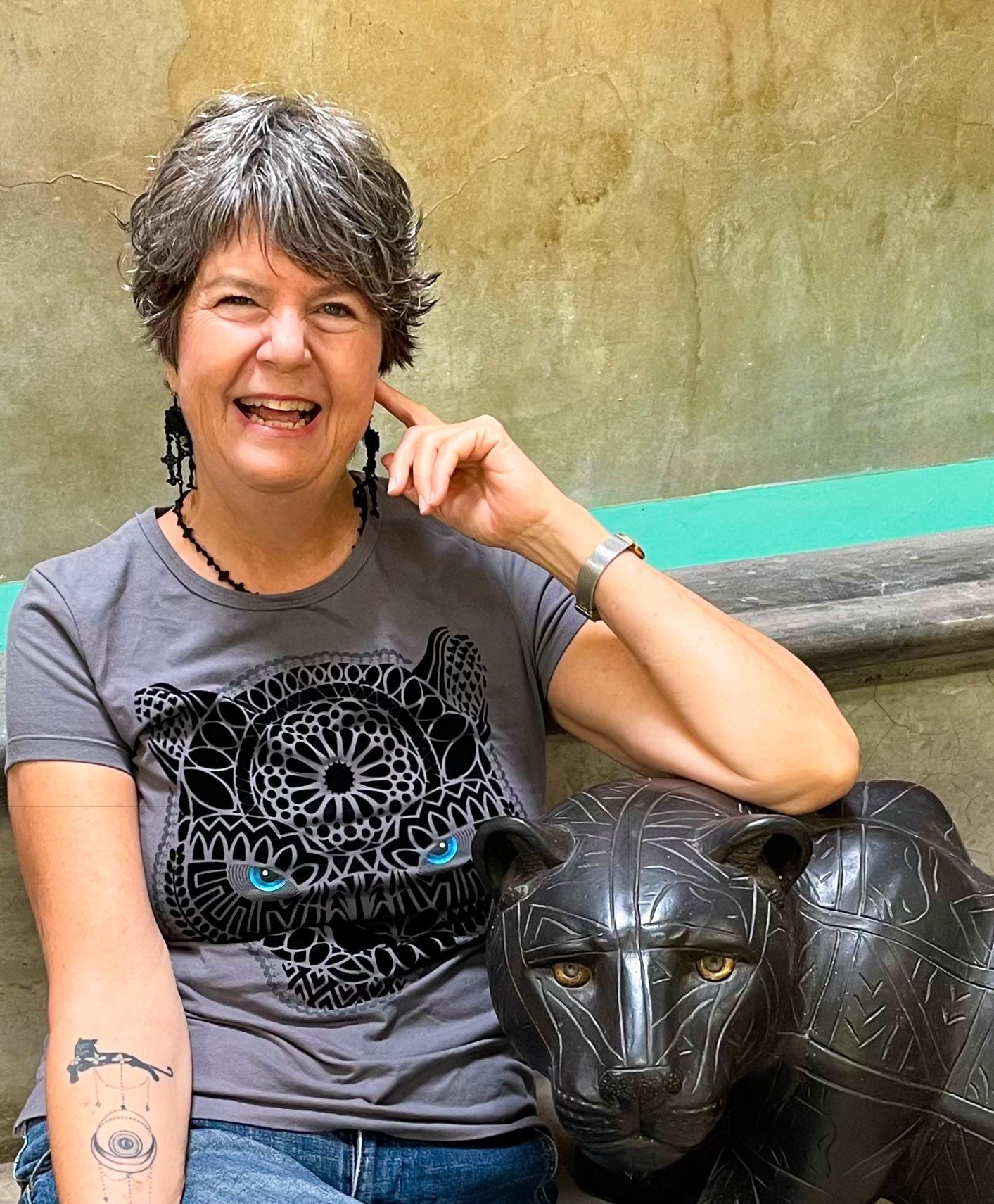 Woman with gray hair wearing headphones and turquoise top, smiling in front of a bookshelf and microphone.