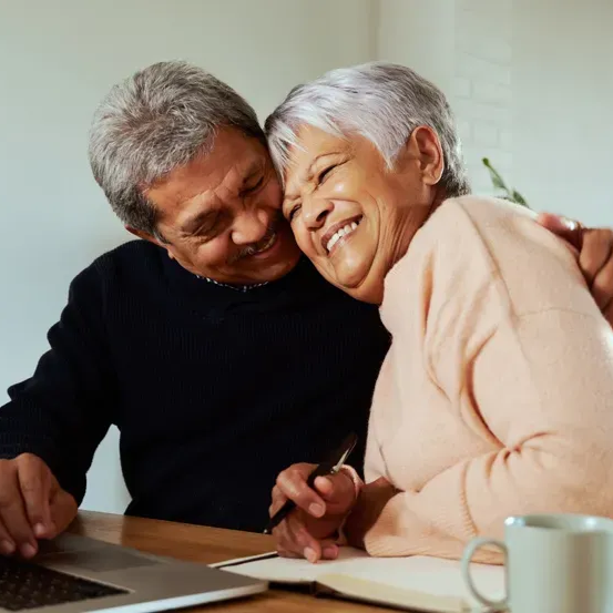 Smiling senior couple embracing, looking at a laptop and writing at a table.