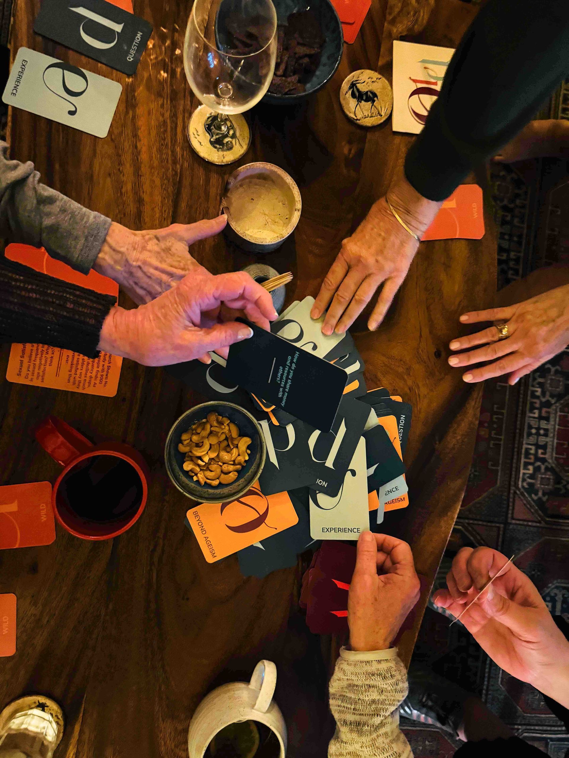People playing a card game around a wooden table, reaching for cards and snacks.