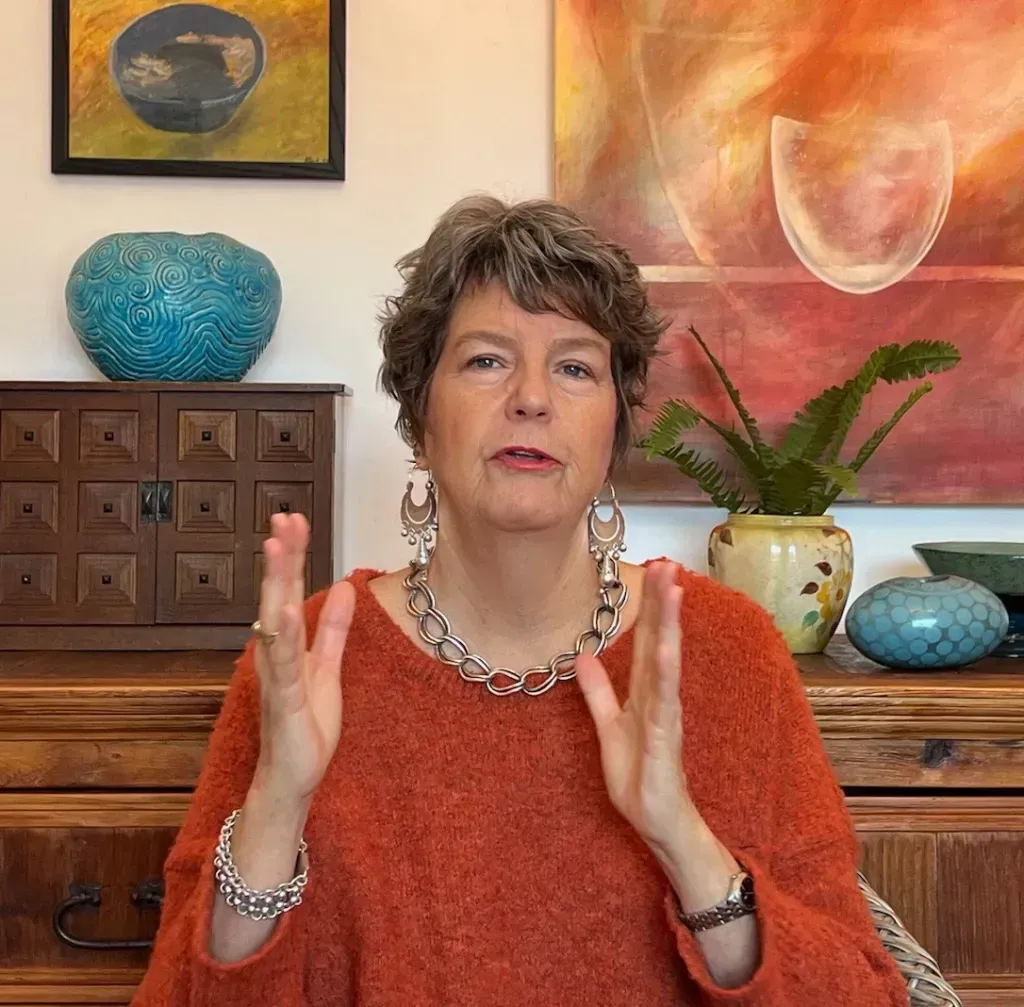 Woman with short gray hair, hands raised, wearing orange sweater and jewelry, in front of artwork and decor.