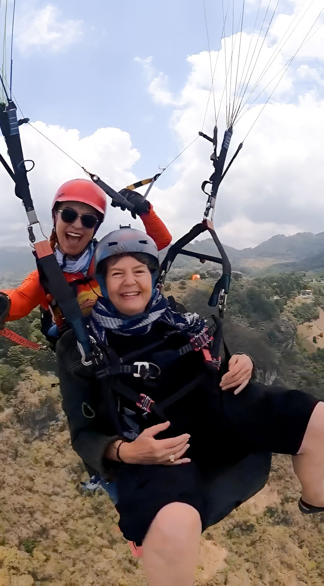 Two people smiling while paragliding over a landscape. One person is piloting.