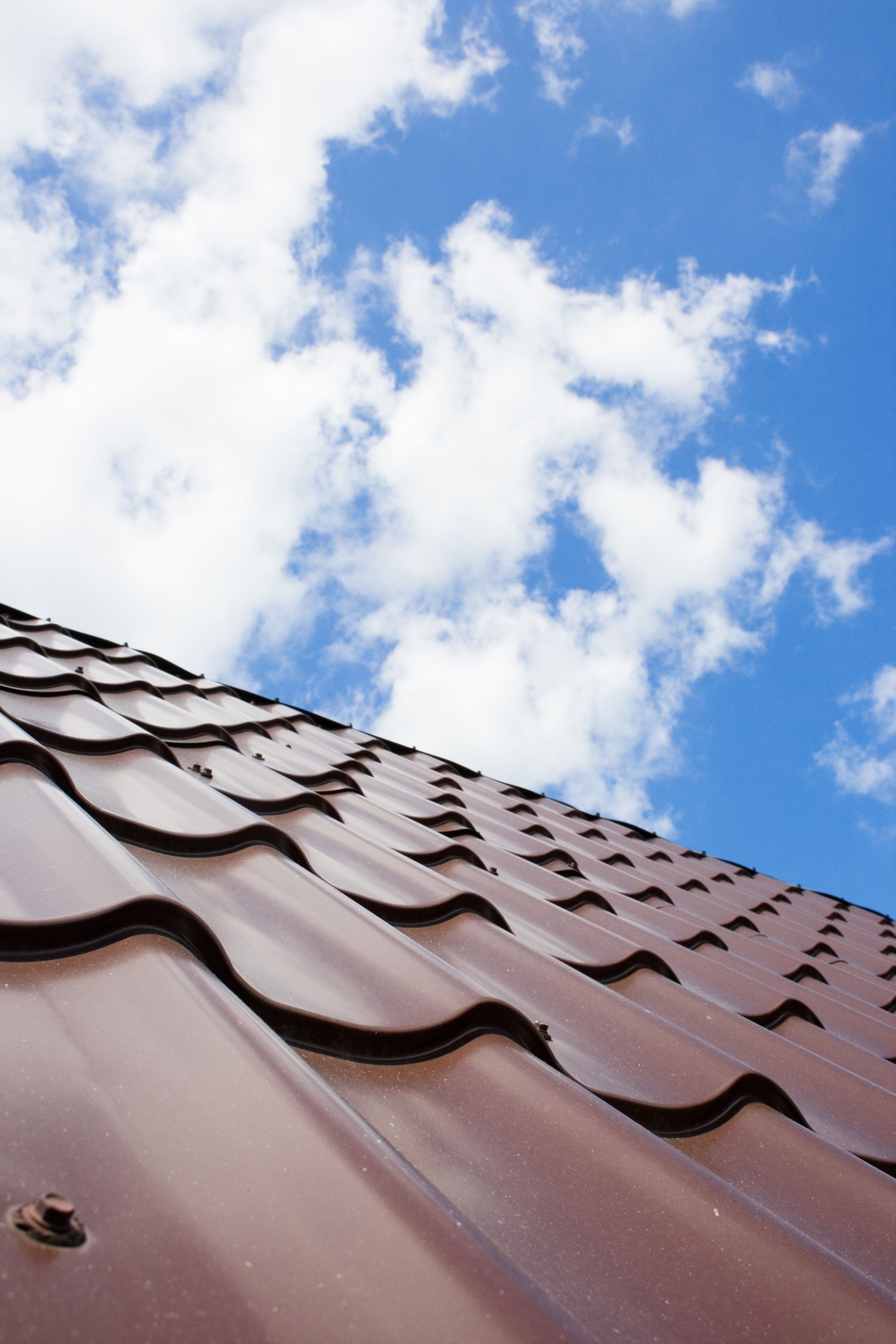 Brown metal roof tiles against a blue sky with fluffy white clouds. Brown metal roof tiles against a blue sky with fluffy white clouds.