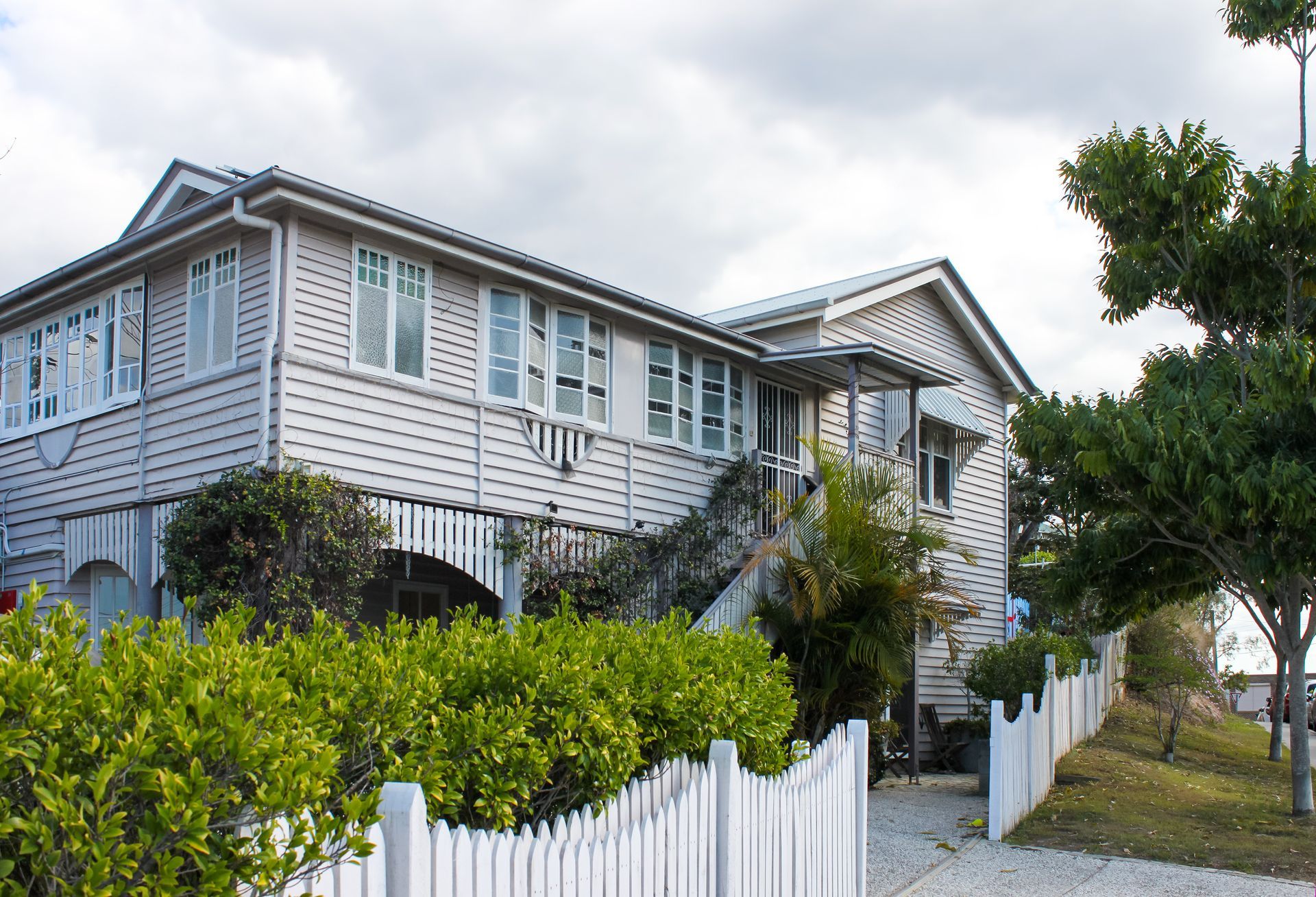 Two-story light-colored house with a white picket fence, steps, and greenery in front. Cloudy sky above.