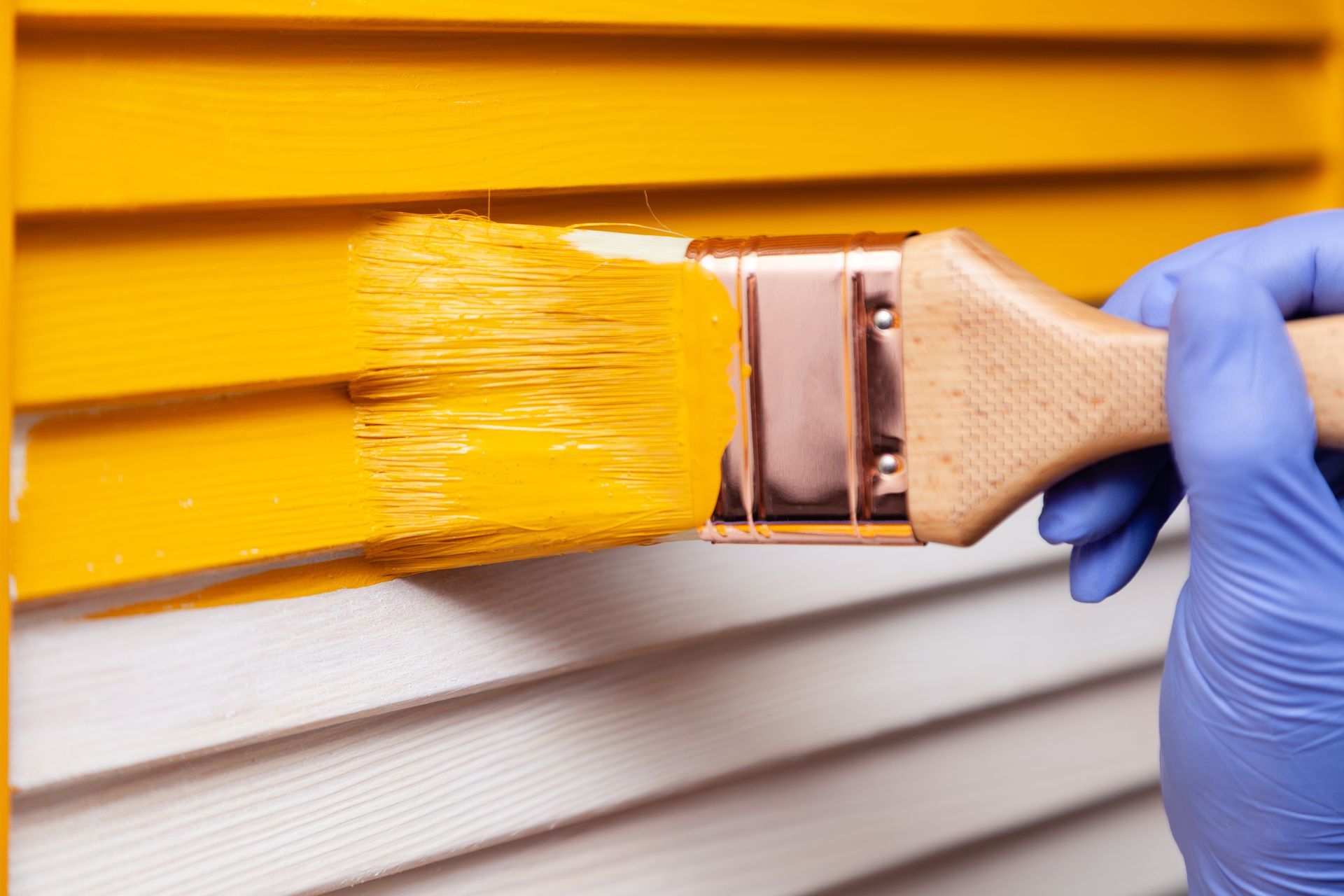 A gloved hand uses a paintbrush to apply yellow paint to a wooden shutter.