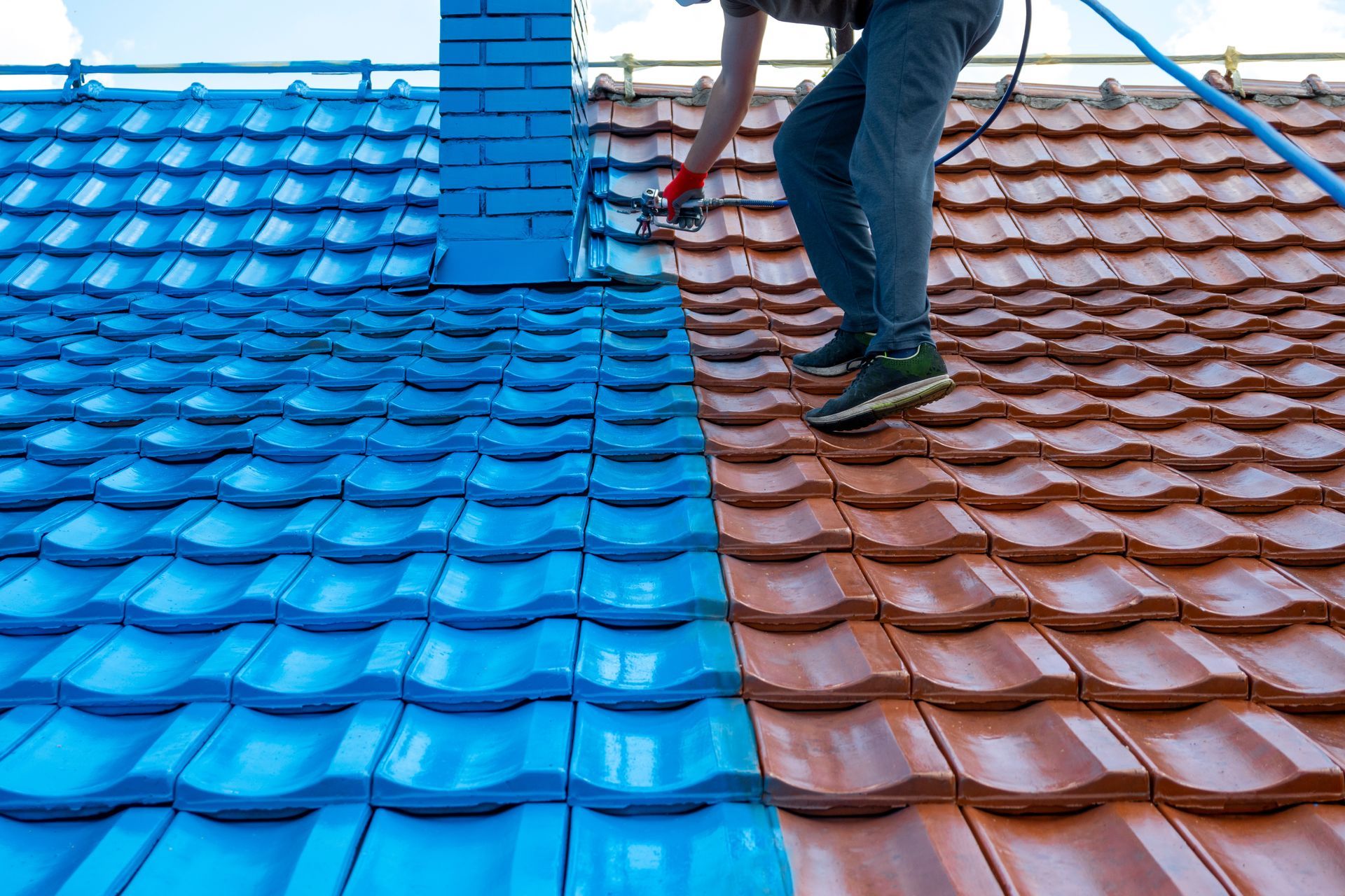 Person spraying blue paint on a red-tiled roof, half-painted. Brick chimney is blue. Person spraying blue paint on a red-tiled roof, half-painted. Brick chimney is blue.