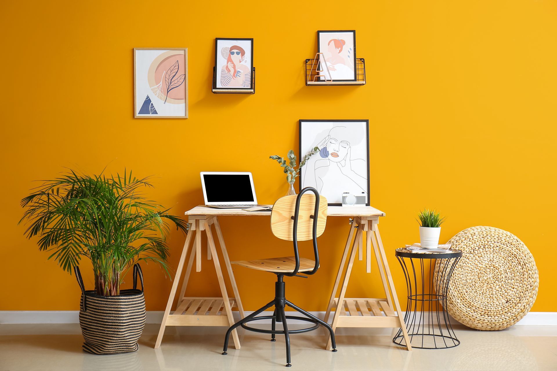 Yellow wall with desk, chair, laptop, art, and potted plants.