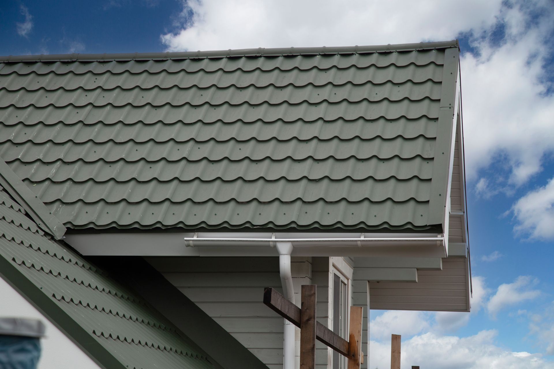 Close-up of a house roof with green metal tiles, white trim, and a clear blue sky with clouds. Close-up of a house roof with green metal tiles, white trim, and a clear blue sky with clouds.