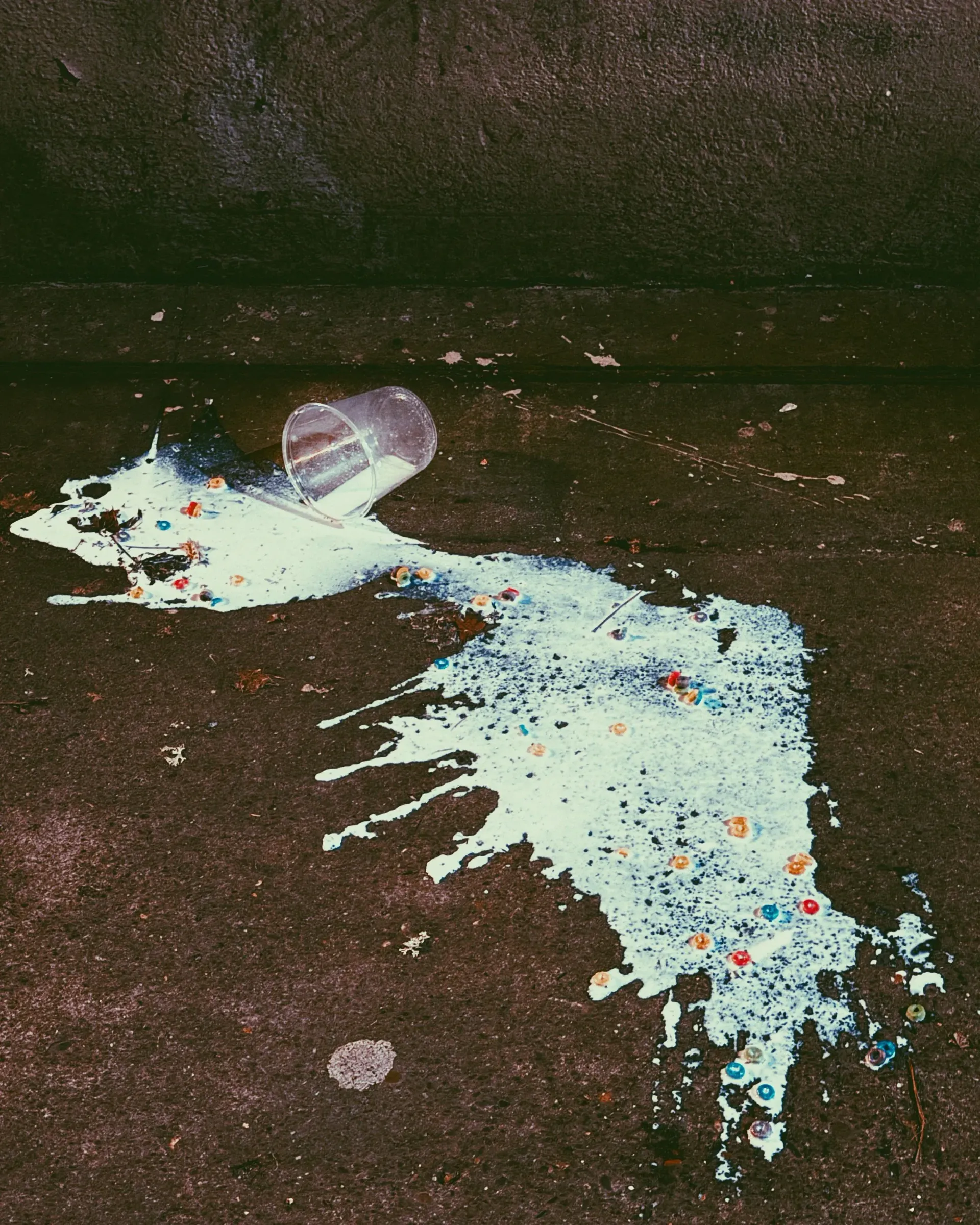 Spilled milk with colorful candies on dark pavement, next to an overturned plastic cup.
