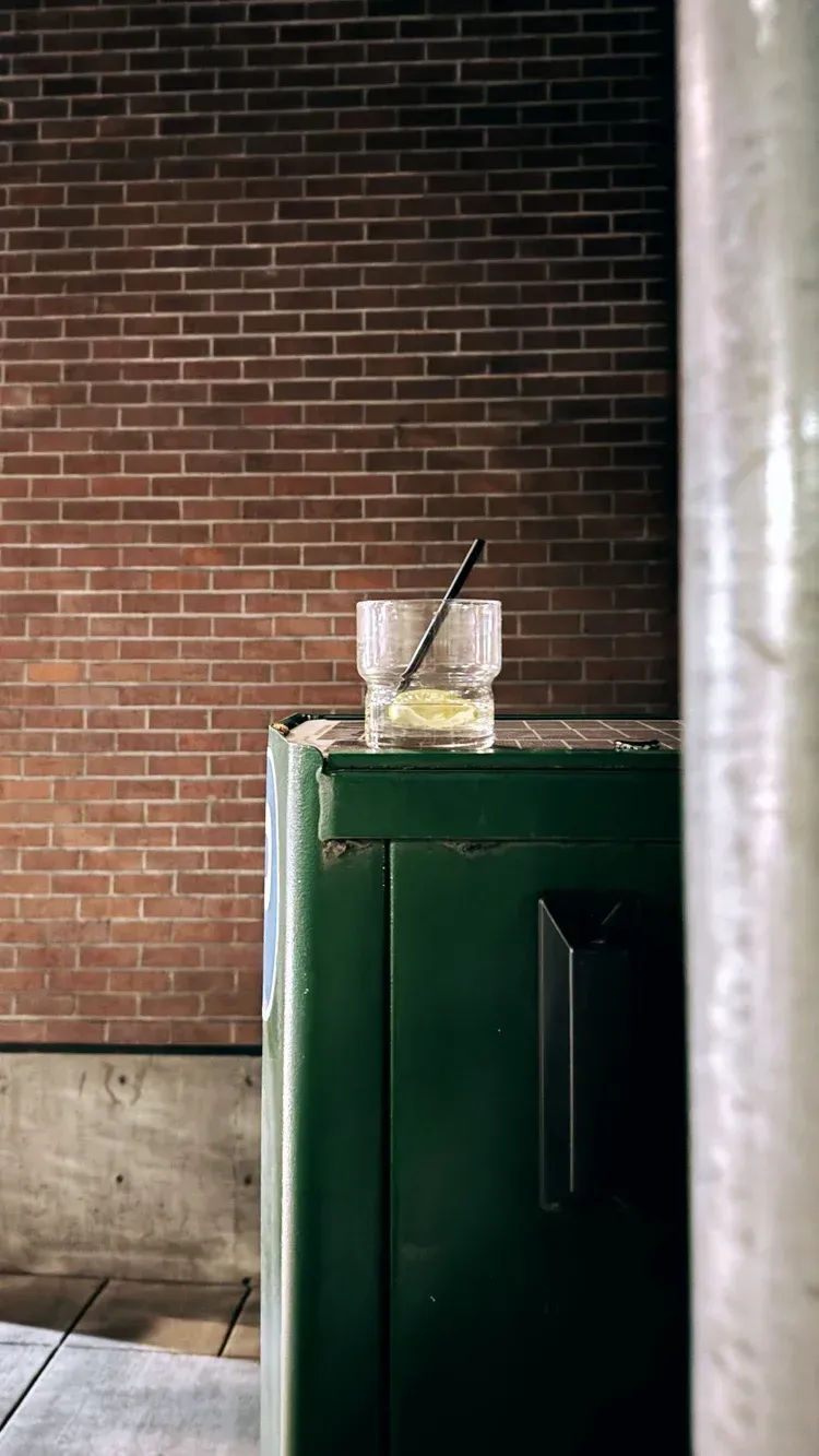 Cocktail glass with straw on a green metal box, brick wall background.