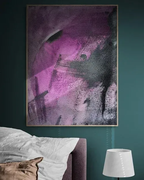 Abstract purple and black painting in a frame hanging above a bed and lamp against a teal wall.