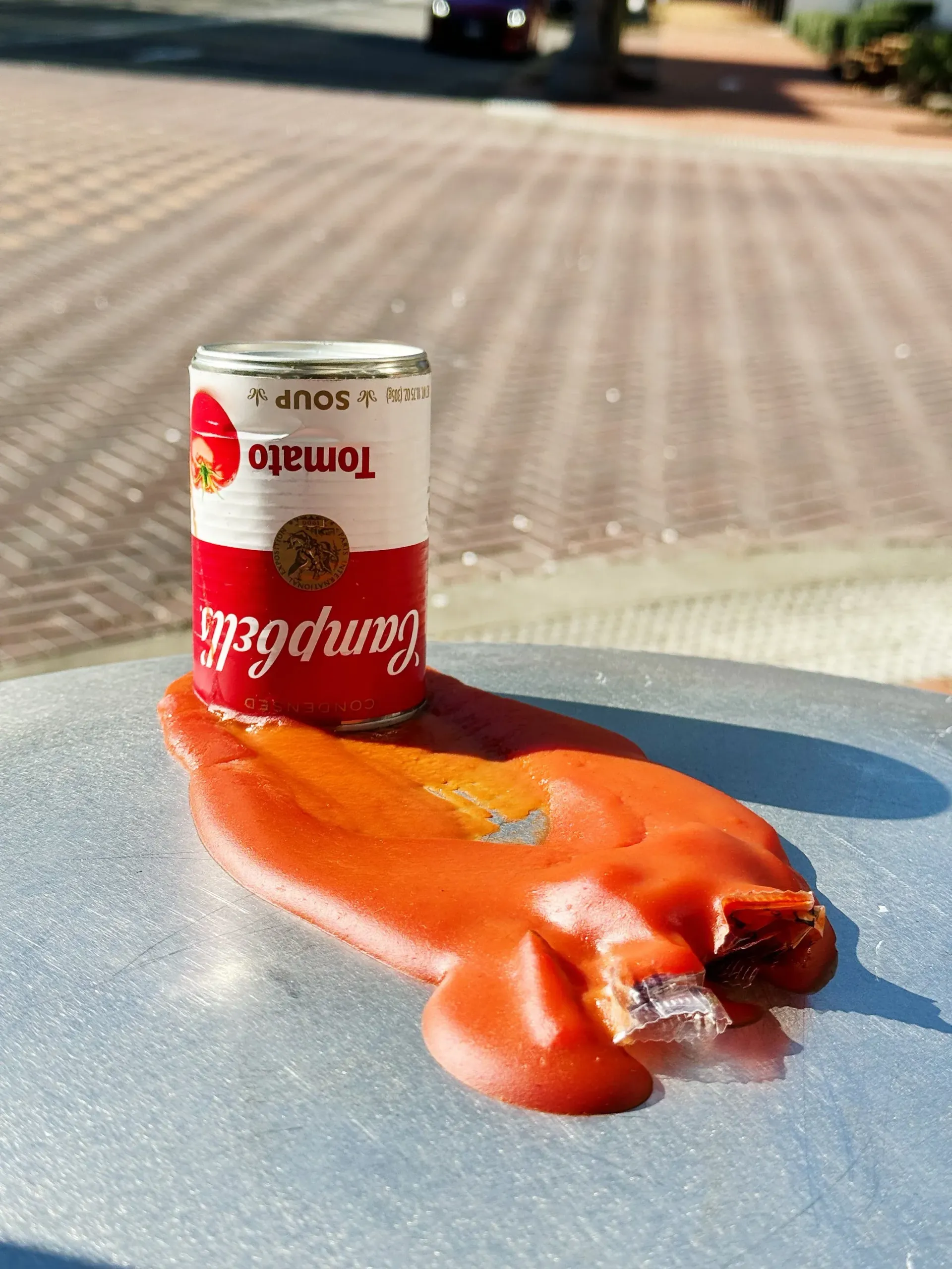 Campbell's tomato soup can on a surface, with spilled soup forming a puddle.