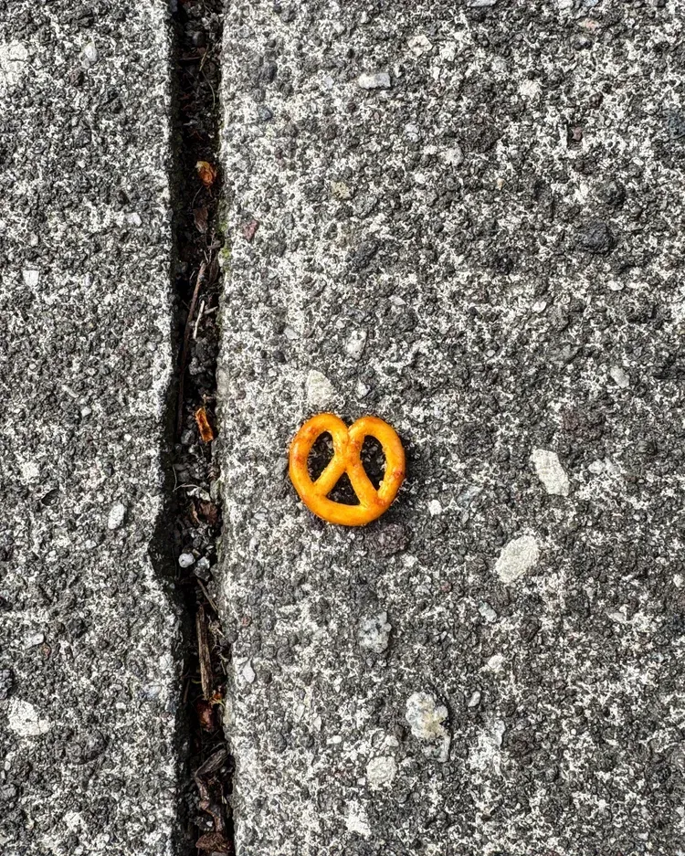 A single small, golden-brown pretzel rests on rough gray pavement beside a dark crack.