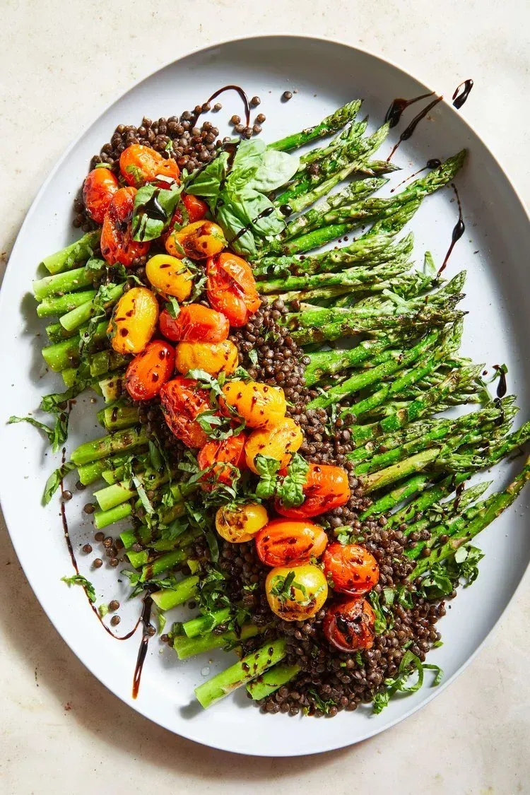 An oval platter with roasted asparagus, lentils, and cherry tomatoes, garnished with fresh herbs and a balsamic drizzle.
