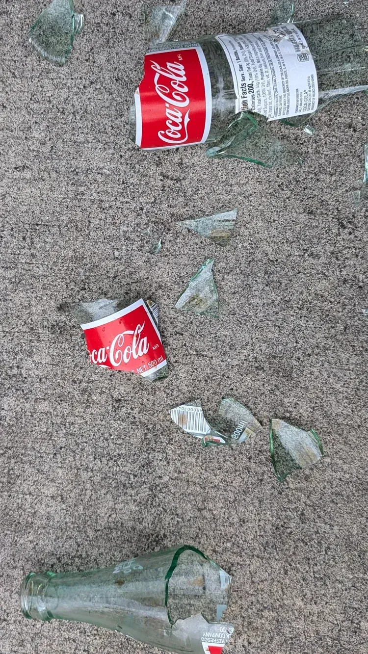 Shattered Coca-Cola bottle and can debris on gray ground.