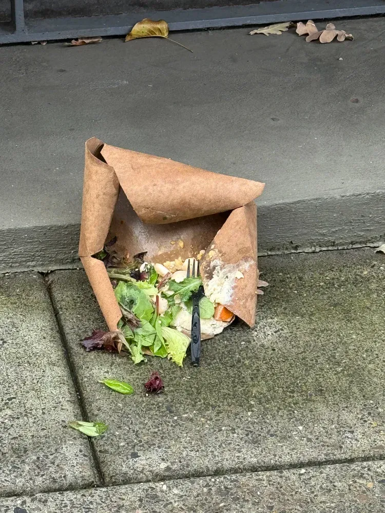 Brown paper takeout container on a sidewalk, spilled contents visible: salad, fork.