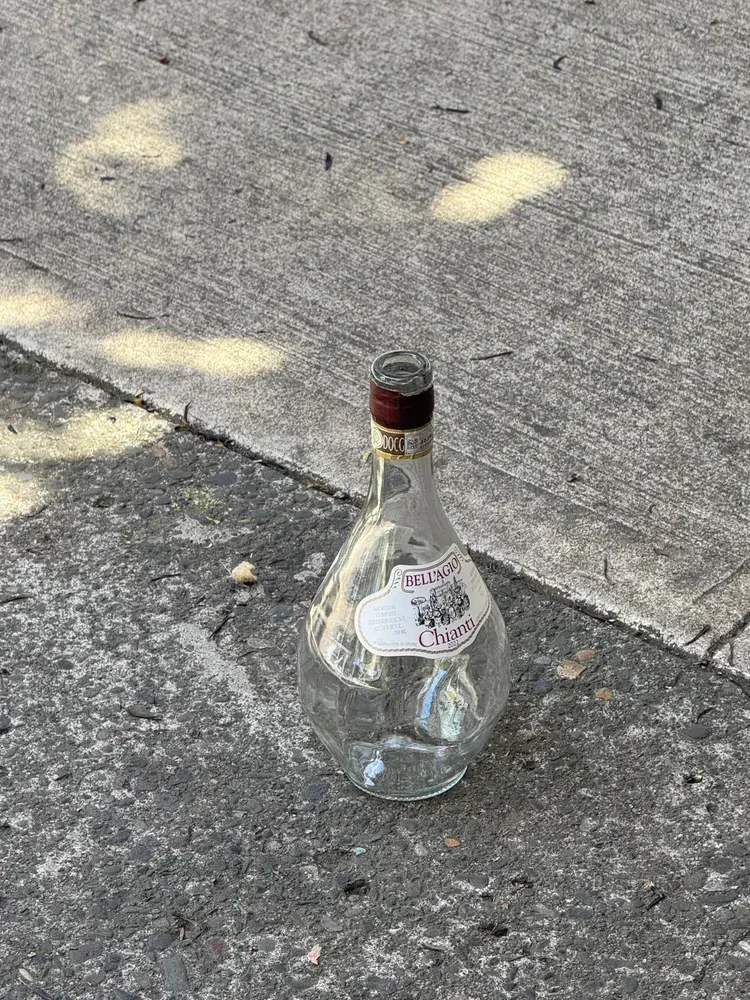 Empty, clear, teardrop-shaped glass bottle with a brown cork and label on cracked concrete.