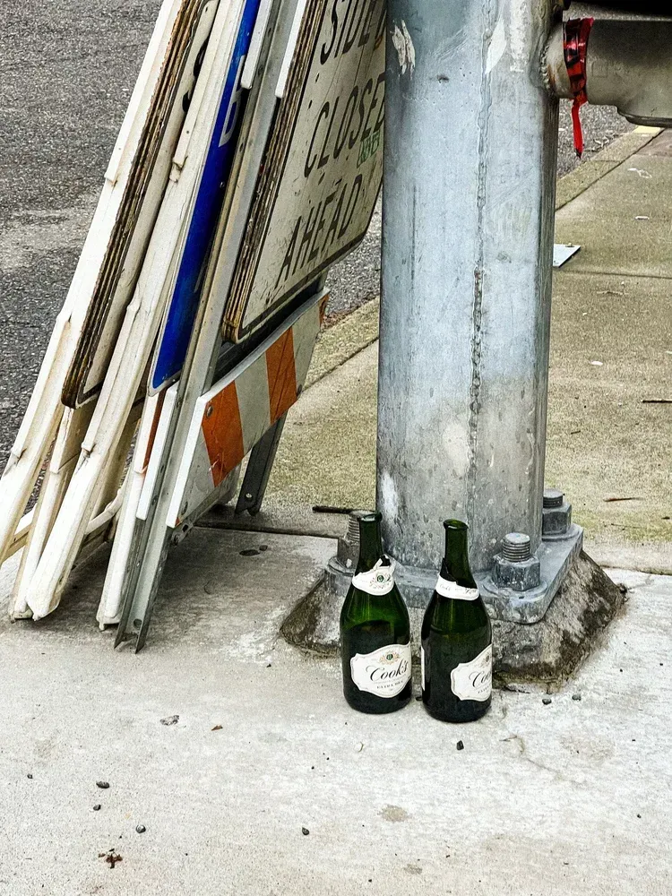 Two empty champagne bottles next to a metal pole, near stacked road signs on concrete.