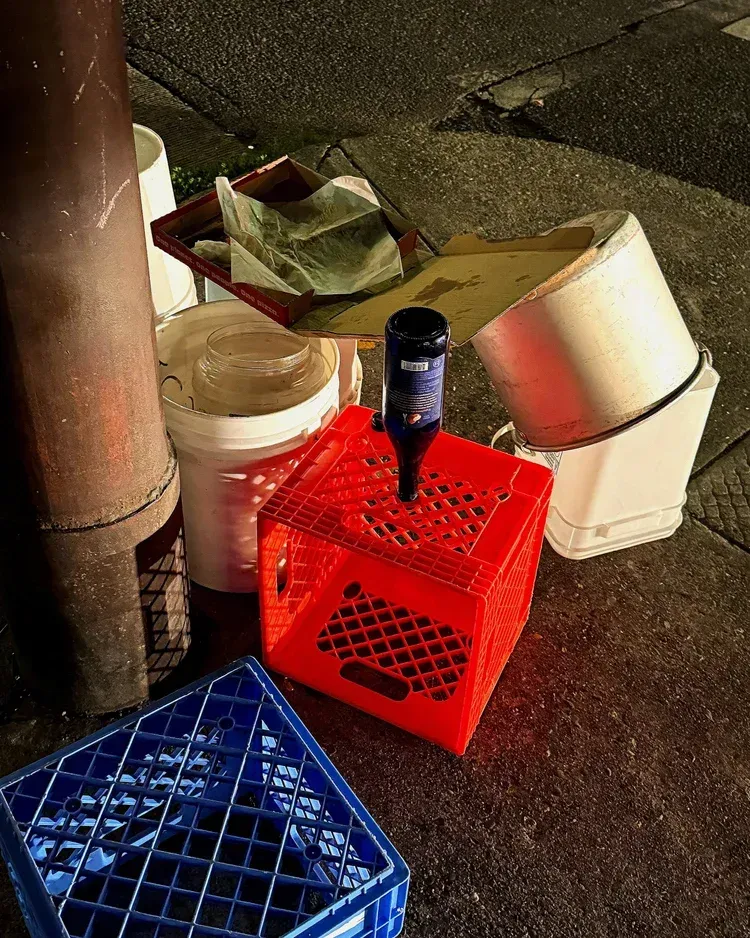 Red crate with a bottle stuck in it, surrounded by trash and other containers on a sidewalk.