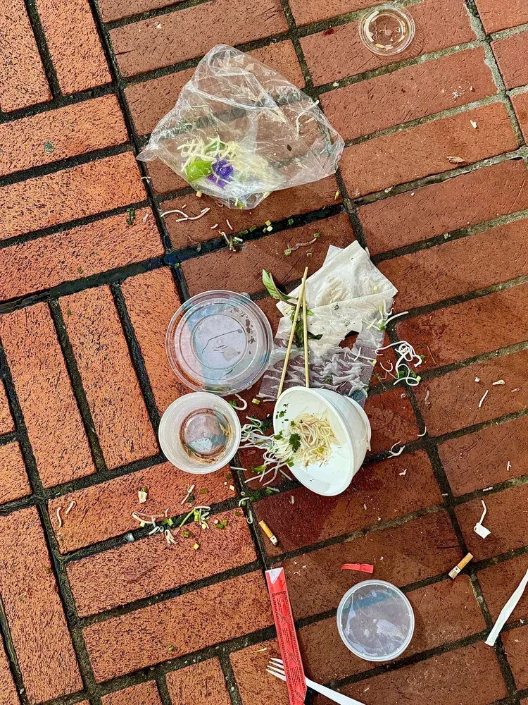 Takeout food waste, including containers, plastic bag, and chopsticks, scattered on brick pavement.